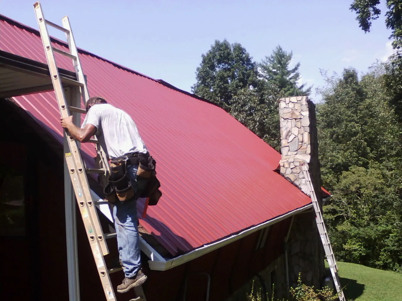 Roofer on a ladder beside a red metal roof and stone chimney — Soffit & Fascia Repair by Roofing Brothers in West College Corner