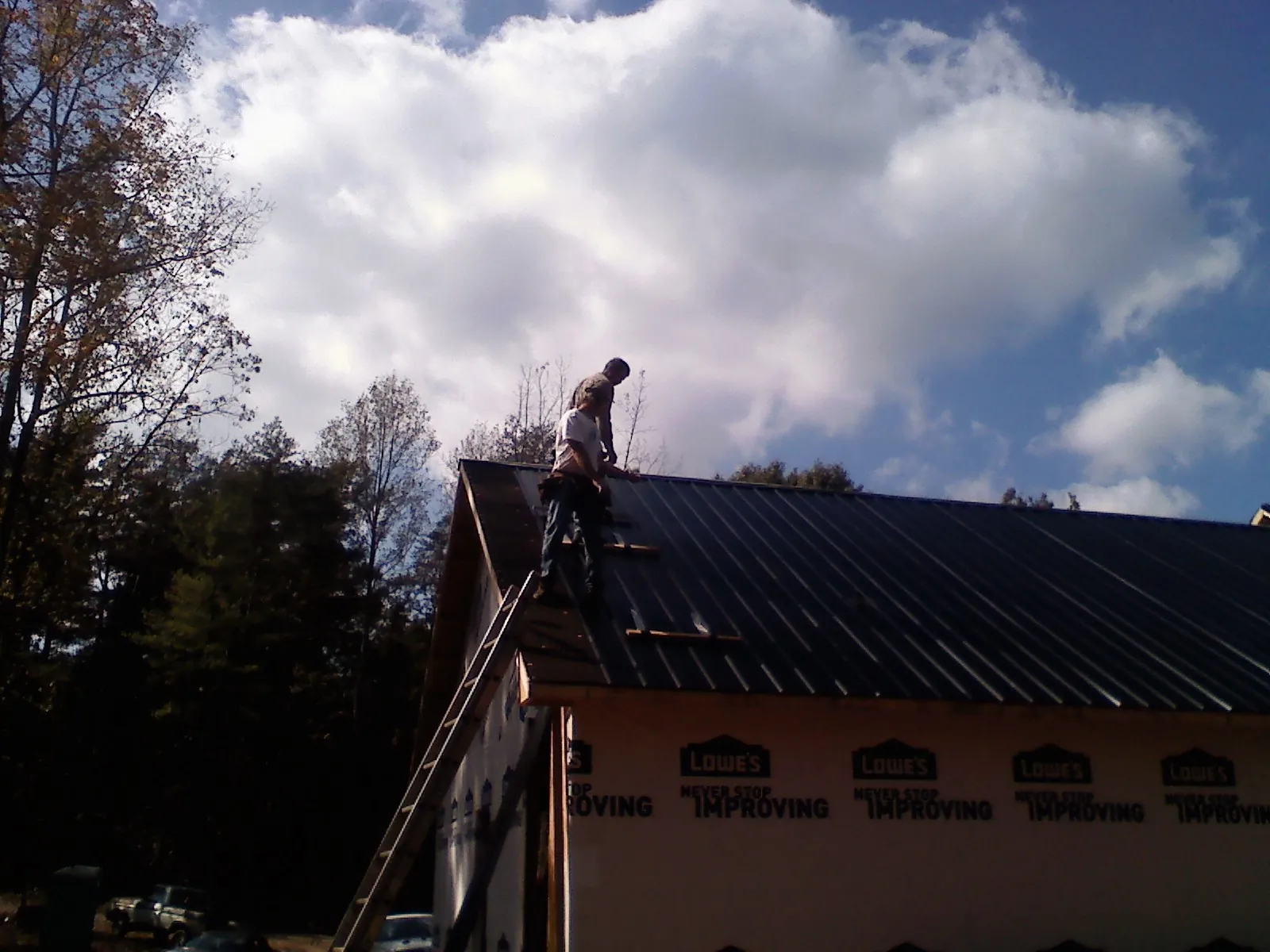 Roofer fastening dark metal panels high against a cloudy sky, from a Roof Inspection job in West College Corner